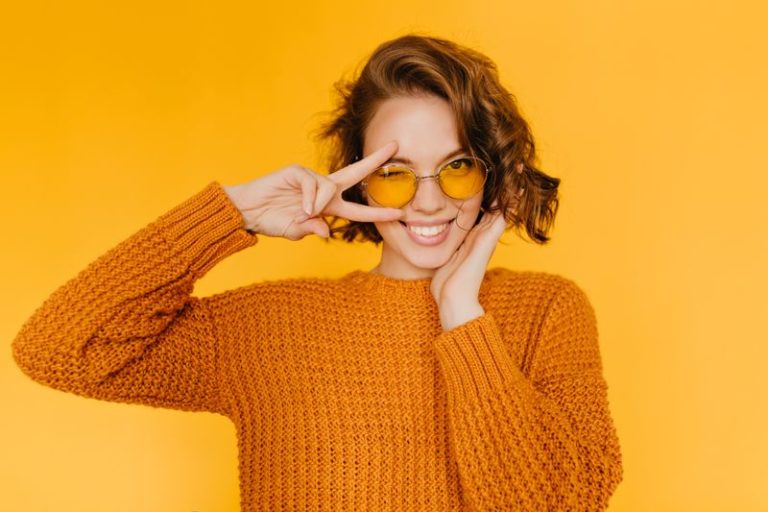 Joyful european girl with shiny curls laughing and showing peace sign on yellow background. Indoor portrait of beautiful lady in glasses and knitted clothes having fun in studio.
