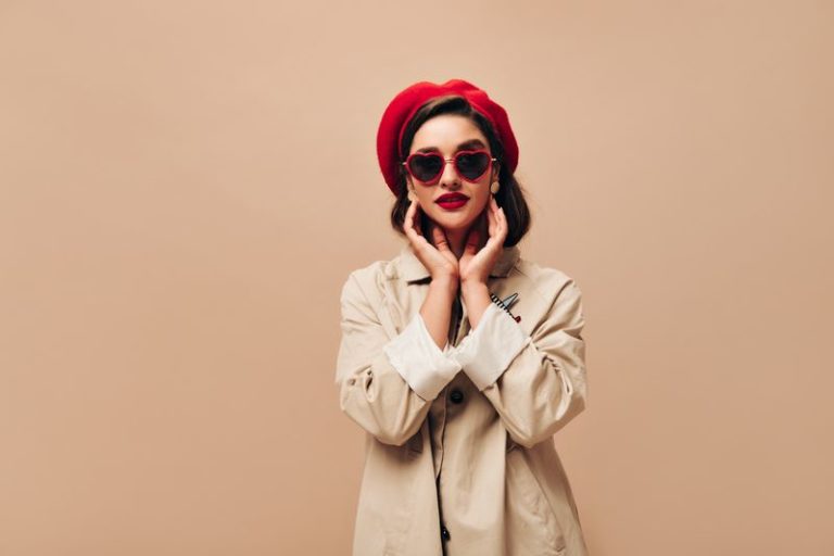 Attractive lady in sunglasses and red beret poses on beige background. Wonderful girl in autumn light coat and bright hat looks at camera