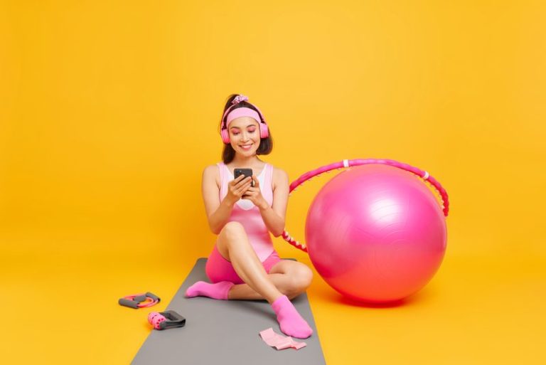 Positive Asian woman with dark hair checks her sport achievements on smartphone application sits on fitness mat dressed in activewear uses swiss ball hula hoop goes in for sport poses indoor