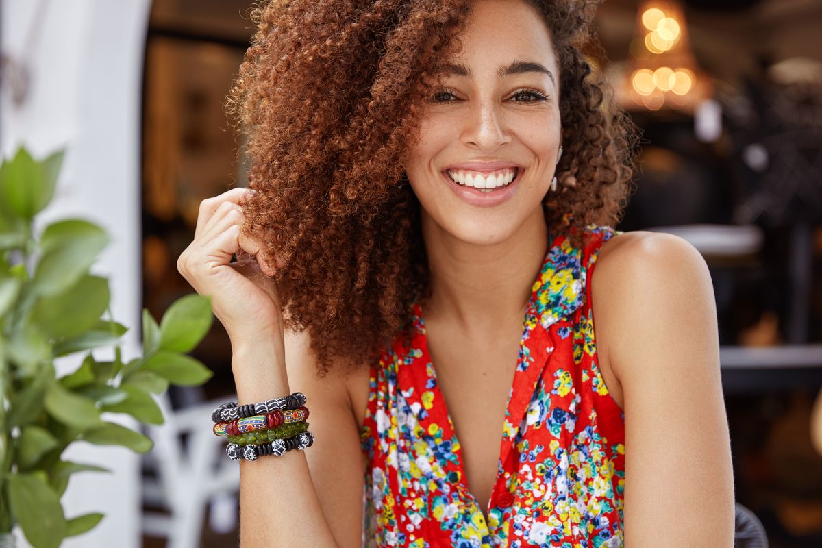 Portrait of good looking happy dark skinned female with curly hair and shining broad smile, demonstrates positive emotions, wears stylish bright blouse. People, ethnicity and beauty concept
